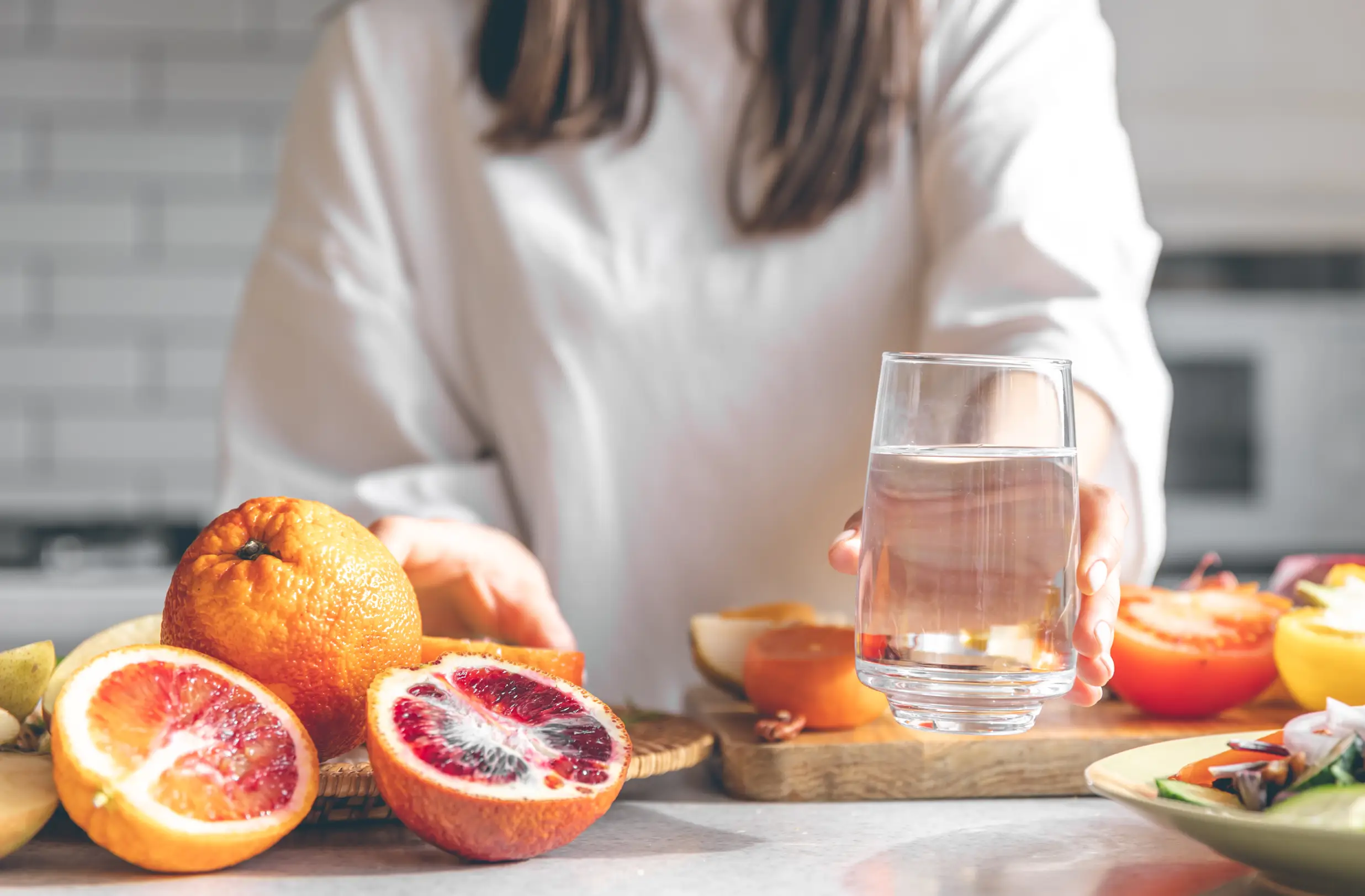 Woman holding a glass of water beside fresh citrus fruits and vegetables on a kitchen counter - iv hydration