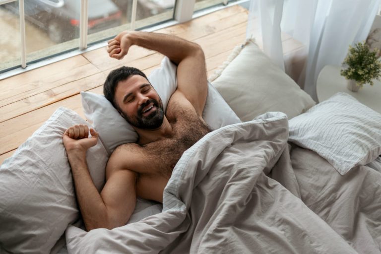 A sleeping man stretching and smiling in bed under a blanket in a bright, cozy bedroom during the morning.